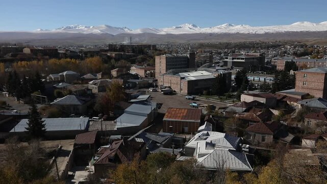 Panoramic view of Gyumri, Armenia, featuring traditional architecture. Majestic snow-capped peaks of Mount Aragats rise in the background under a clear blue sky during a sunny autumn day.

