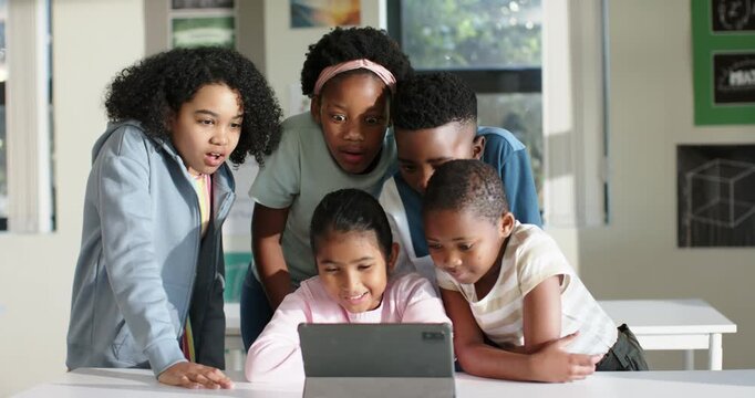 Diverse child students crowding seated peer using tablet in classroom reacting to surprise laughing