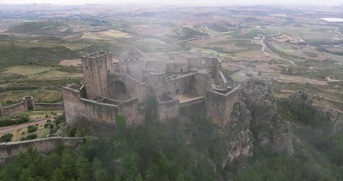 Wide aerial drone shot of Castillo de Loarre on a rocky outcrop in the Pyrenees, Spain, medieval fortress surrounded by fog and dramatic cloud shadows over the landscape