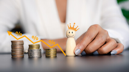 Businesswoman hand holding smiling wooden king doll with decreasing coin stacks and percentage graph for cost reduction concept.