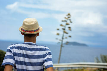 Teenage boy sitting on a coastal path above the sea, observing a flowering agave tree. Peaceful summer moment, reflection, and connection with nature, ideal for travel and lifestyle concepts.