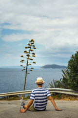 Teenage boy sitting on a coastal path above the sea, observing a flowering agave tree. Peaceful summer moment, reflection, and connection with nature, ideal for travel and lifestyle concepts.