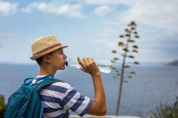 Teenage boy drinking water from a plastic bottle during a summer hike with sea view and a flowering agave in the background. Outdoor adventure, hydration, and scenic coastal exploration.