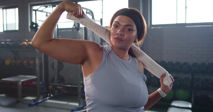 Woman in sleeveless top holding towel behind neck, flexing bicep in gym, checking results on watch