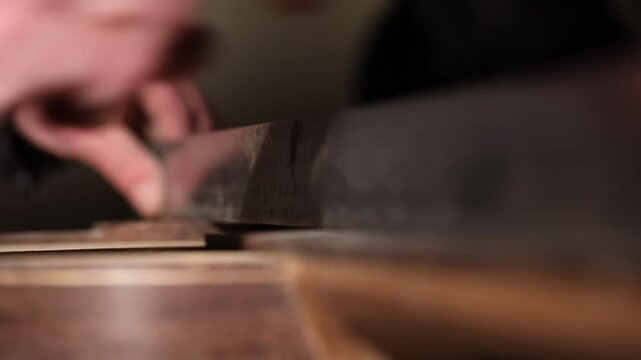 A close-up shot of a craftsman's hands sanding the rough edge of a wooden structure. He uses a small wooden tool to smooth the surface in a dim workshop.

