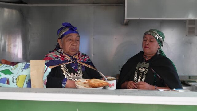 Two mapuche women in traditional clothing enjoying a meal of sopaipillas and mate in a food stall