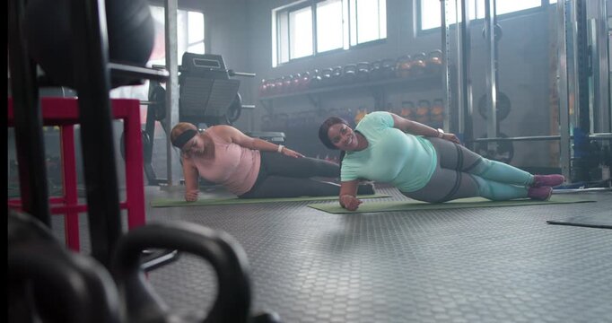 Diverse female friends on left forearms holding side plank on gym mats, shifting hips engaging core