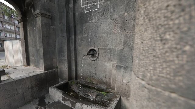 Zoom out revealing a traditional Armenian stone drinking fountain known as Pulpulak. The grey volcanic tuff structure features arches and columns situated outdoors on a sunny day.

