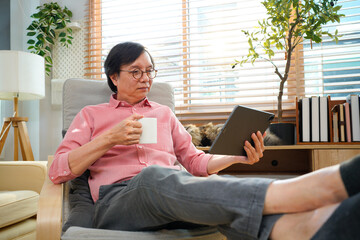 A smart mature man with coffee mug looks in a tablet in his hands while sitting in a comfortable chair in the living room