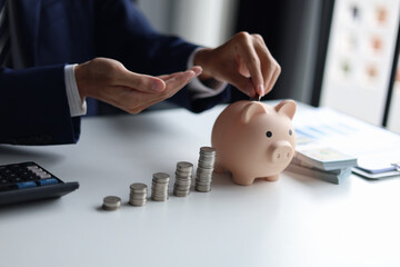 A&nbsp;person in a suit putting a coin into a piggy bank with stacks of coins and money on the table. Saving money and financial planning, financial growth, investment, and future planning concept.