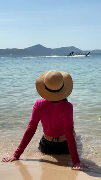 Young woman in bright clothes and straw hat sitting on the beach by the azure sea, enjoying the stunning view and gentle waves.	
