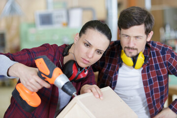 happy female carpenter drilling wood while colleague working in background