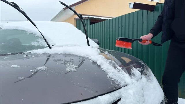 Individual using a snow brush to clear snow from the hood of a car, showcasing the action of snow removal, with a green fence and building in the background