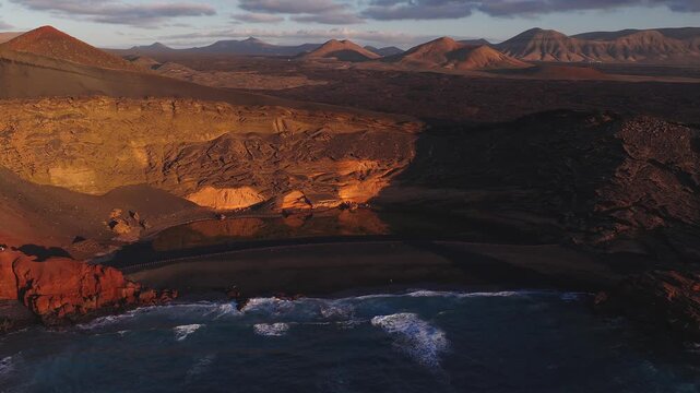 Aerial view of El Golfo and Charco de los Clicos in Lanzarote, with black sand, crater lagoon, rust red cliffs, gentle waves, tiny visitors, and Timanfaya peaks at golden hour.