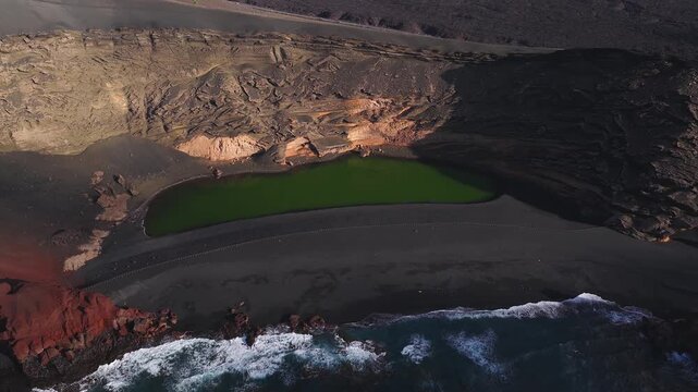 Aerial drone sweeps reveal Charco de los Clicos at El Golfo, Lanzarote, emerald lake, black sand, rust red cliffs, waves crash, coastal road, long afternoon shadows.