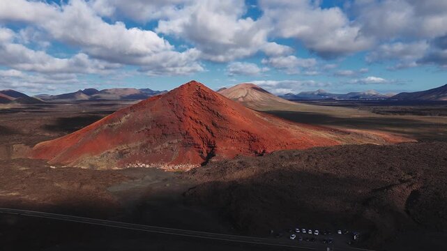 Drone sweeps over Lanzarote, Canary Islands, a red cinder cone rises from black lava fields, textured stratified slopes, tiny cars in a parking area, patchy clouds move