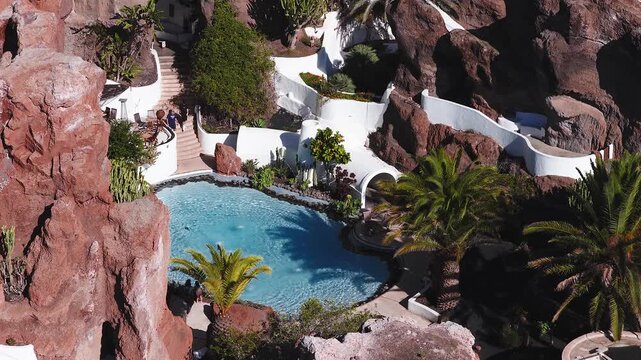 Aerial view of Lagomar house in Lanzarote, Canary Islands. Jagged volcanic cliffs frame a turquoise circular pool, white terraces, arched tunnels, and palm trees.
