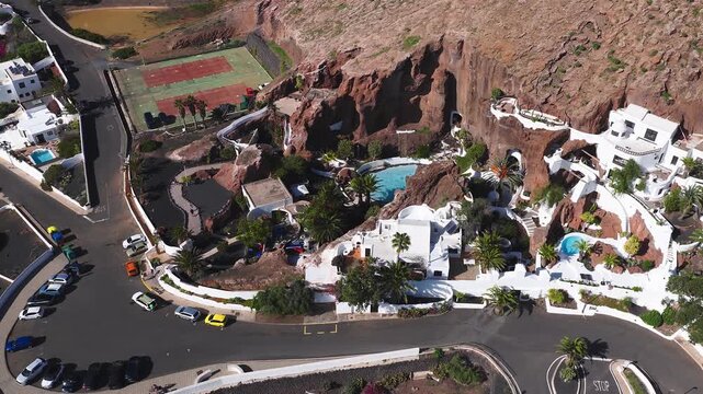 Aerial daytime view of Lagomar Casa Museo in Lanzarote shows carved caverns, winding terraces, circular pools, tennis courts, parked cars, and a few pedestrians.