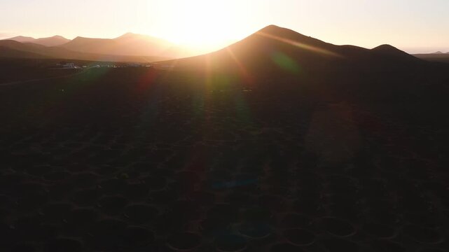 Aerial view of La Geria vine pits, a silhouetted volcanic cone, and layered ridges in Lanzarote, Canary Islands. The camera sweeps with strong backlighting and lens flares.