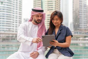 Business professionals, Middle Eastern and Asian discuss ideas using a tablet while sitting in a modern city with tall buildings.