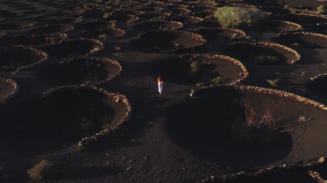 Aerial La Geria in Lanzarote, Canary Islands, shows zocos in volcanic ash as a red haired woman in white walks among rings. Warm low light casts long shadows.