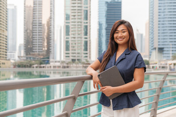 Asian businesswoman stands by a railing in a smart city, using a tablet while embracing urban life and digital work culture.