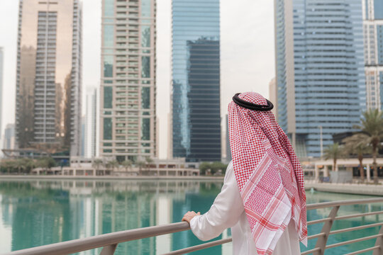 Middle Eastern businessman in traditional attire stands in an urban area looking at skyscrapers. He represents success in finance and real estate.