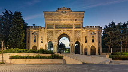 Fototapeta premium Istanbul University's historic main gate, illuminated at dusk, with ornate architecture and clocks. Istanbul, Turkey.