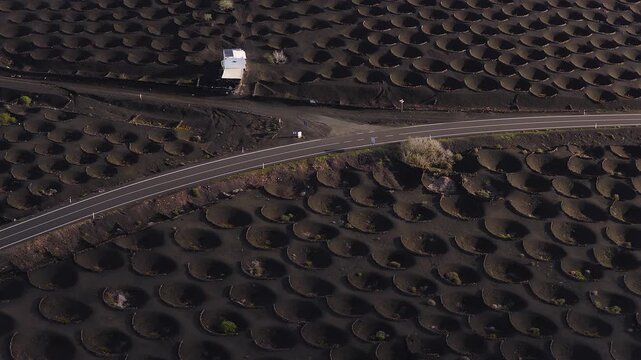 Aerial view of La Geria vineyards in Lanzarote shows zocos in black lapilli, a curving road, and a small white building, with low warm sunlight and long shadows.