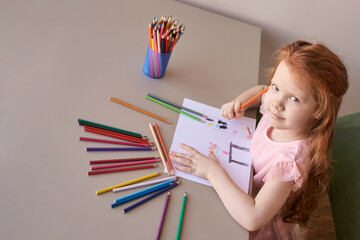 Young caucasian girl drawing with colorful pencils on table in bright room.