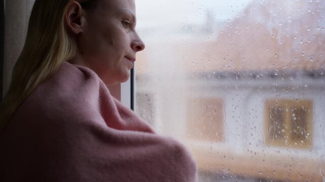 Pensive woman wrapped in warm blanket feeling lonely and sad while look at rain outside window, dealing with feelings of depression and solitude on cold autumn day