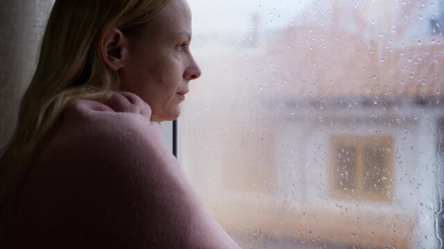 Pensive blonde woman in contemplation, wrapped in pink blanket, gazing out window on cold, rainy day, concept emotions of solitude, melancholy, and inner battles with mental wellbeing