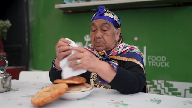 Elderly mapuche woman in traditional clothing eating fried sopaipillas at a food truck in chile