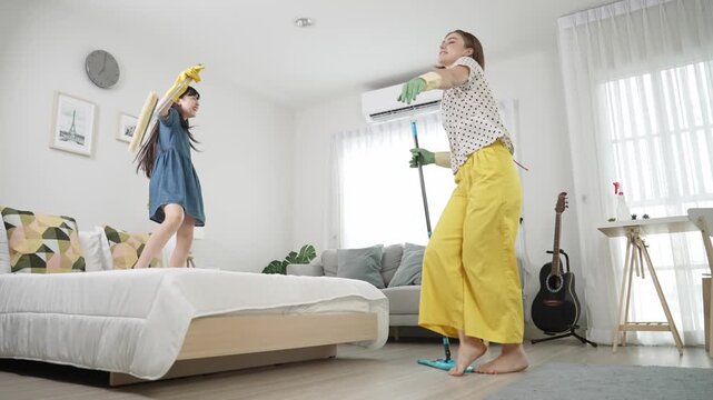Energetic child jump on bed and attractive mom dance while cleaning bedroom. Happy mom and girl doing housework together. Attractive daughter listening and moving to music with lively mood. Pedagogy.