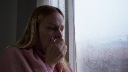Sick woman with cold or flu coughing and drink hot tea from mug while look out window on rainy day. Sad and lonely person dealing with illness and mental health struggles at home
