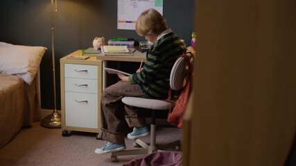 Full slowmo shot of focused blond Caucasian student boy sitting on rolling computer chair at well-organized study desk and concentrating on school assignment, demonstrating academic motivation