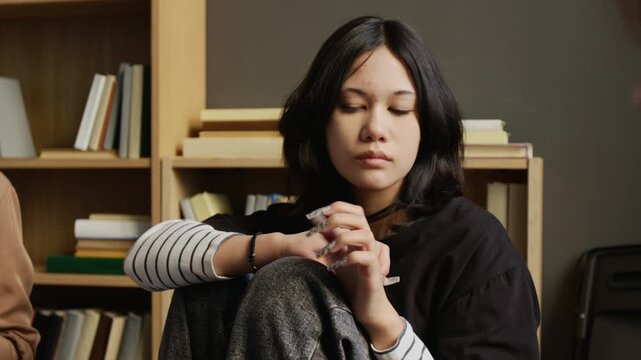Medium shot of emotionally withdrawn teenage girl with black hair fidgeting fingers while during group counselling session in therapy room