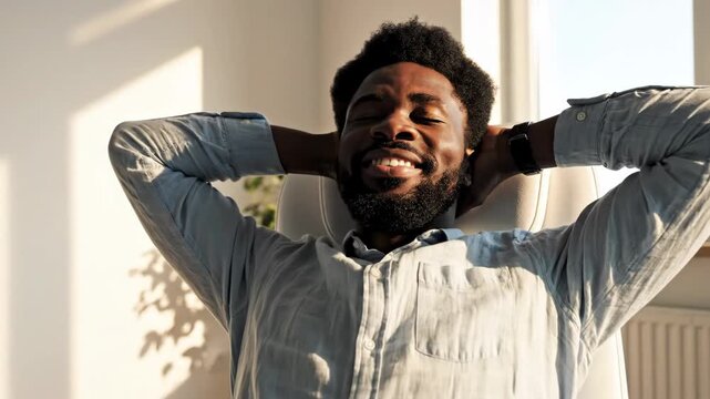 Relaxed Businessman in Office Chair - A smiling African-American man relaxes in an office chair with his hands behind his head. Sunlight streams in from a nearby window, casting shadows on the wall.