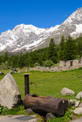 Traditional wooden water fountain in an alpine meadow with green grass, stone walls, forest and snowcapped Mont Blanc mountains near Courmayeur on a sunny day.