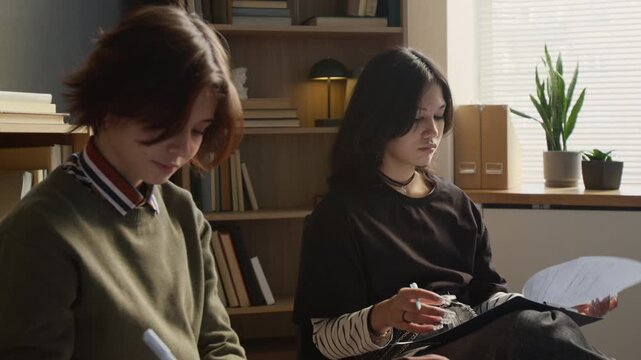 Young teenage girl peeking answers in questionnaire of groupmate while filling out mental health tests on clipboards at therapists office