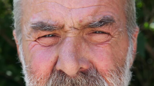 Blue eyes of elderly male blinking and looking into camera outdoor. Wrinkled face of senior human with a sad sight. Upset facial expression of pensioner. Close up