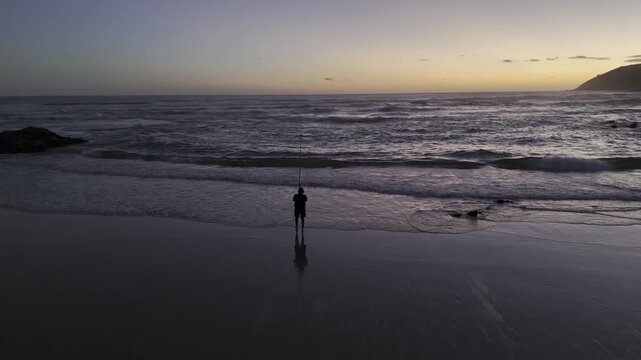 Drone slowly flies backwards as lone fisherman on beach reels in a catch at sunset on the Garden Route in Wilderness, South Africa