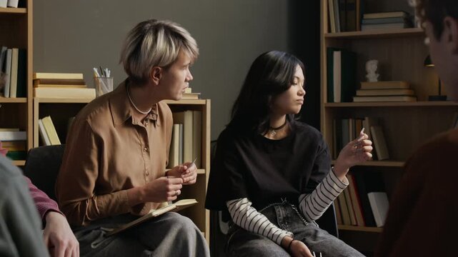 Teenage girl with black hair remaining emotionally withdrawn while adult female psychotherapist talking to adolescent during group discussion in therapy room