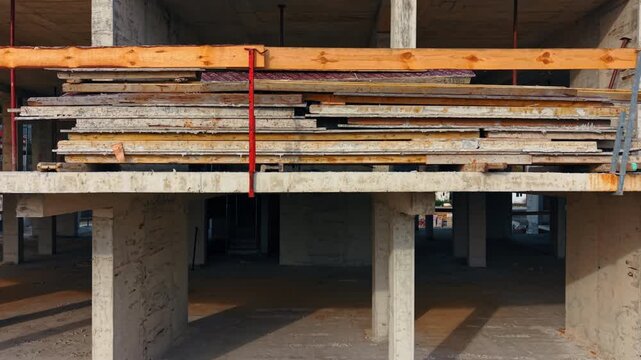 A construction site shows a large stack of building materials resting on beams in an incomplete building. The structure has exposed concrete and columns.