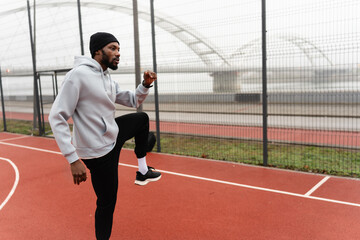 Young Black man warming up with high knees on an outdoor basketball court near a bridge, wearing sportswear on a cloudy day, focused on fitness, cardio training, and urban active lifestyle