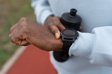 Close-up of a Black athlete checking a smartwatch while holding a water bottle outdoors. Fitness tracking, healthy lifestyle, training routine, focus and motivation