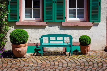 Green Bench Sits in Front of a White Wall and Windows With Green Shutters in a Garden Setting During Bright Daytime