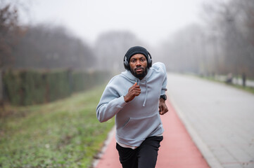 Fototapeta premium Focused Black man running outdoors wearing headphones and a hoodie on an overcast day, expressing determination, energy, and motivation, capturing fitness, endurance, and healthy active lifestyle