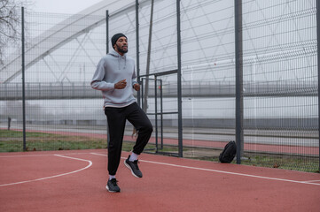 Obraz premium Young Black man warming up with high knees on an outdoor basketball court near a bridge, wearing sportswear on a cloudy day, focused on fitness, cardio training, and urban active lifestyle