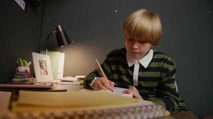 Medium shot of blond Caucasian boy child concentrating on solving math problem or writing assignment, sitting at cozy home study desk surrounded by lots of stationery and educational materials